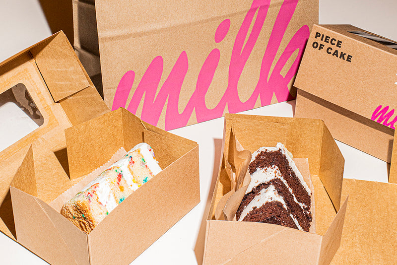 Side view of the Milk Bar Birthday Cake slice and Chocolate Birthday Cake slice in a to-go cardboard box. 
